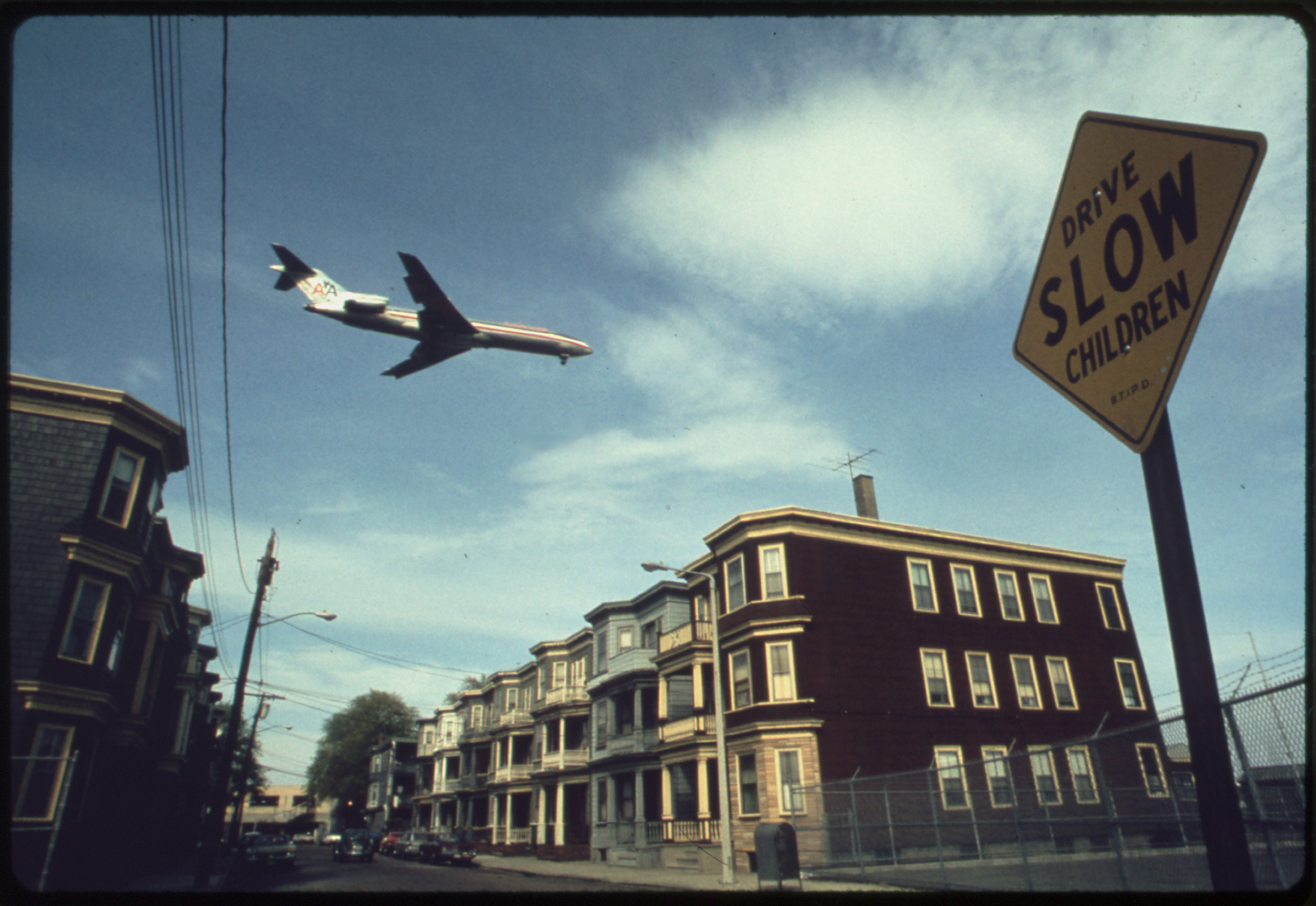 An airplane flies over houses in Boston in 1973.