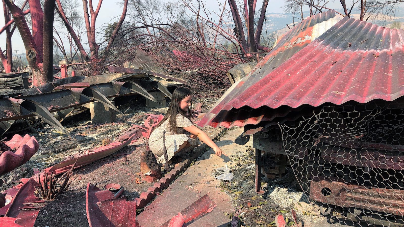 A photo of a girl digging touching the rubble of her burned home in Talent, Oregon. She is in the center of the image in a white outfit. The ground and debris are stained red from an airdrop fire retardant.