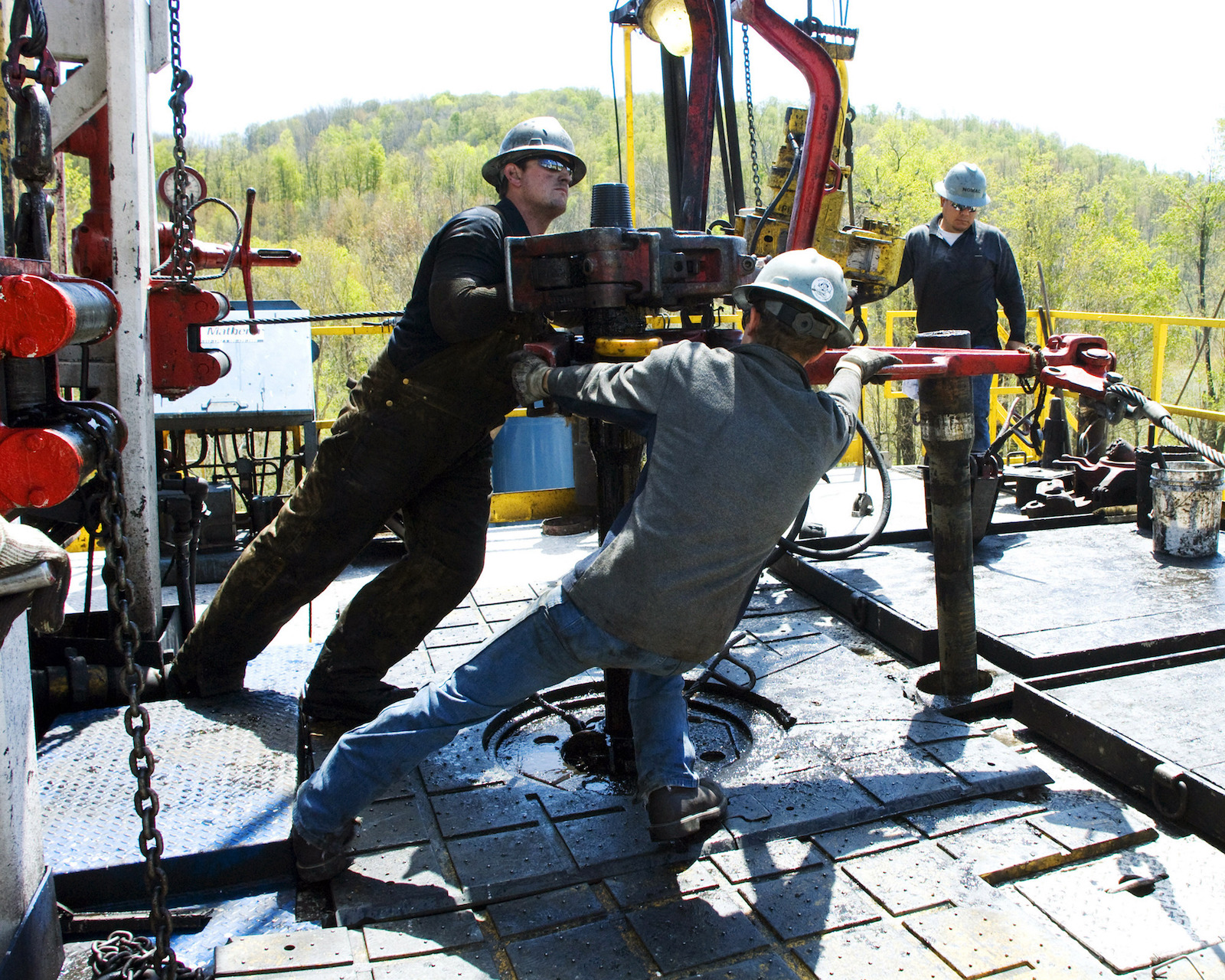 Two men in hard hats are working with a large piece of equipment in the center of the photo. There are green trees in the background.