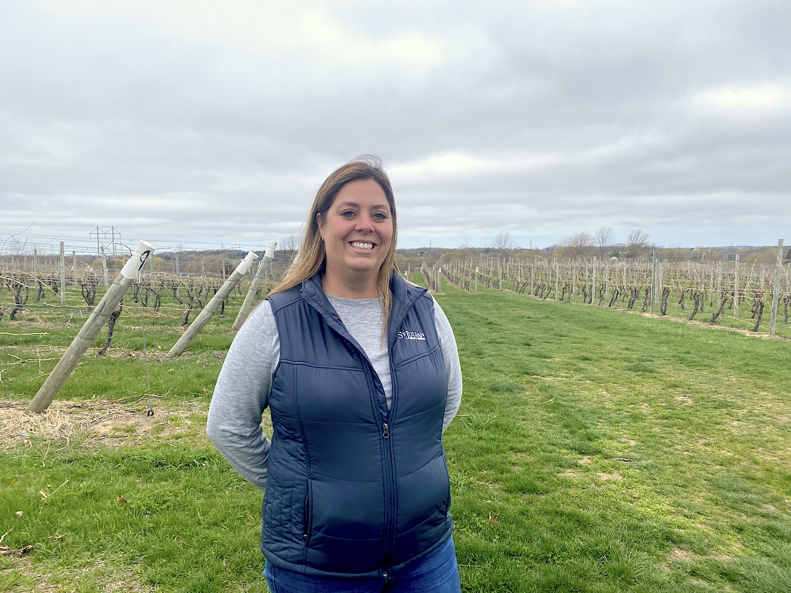 A photo of a woman in a black puffer vest and a long blue-gray shirt. She has blonde hair and is smiling. In the background are green fields.