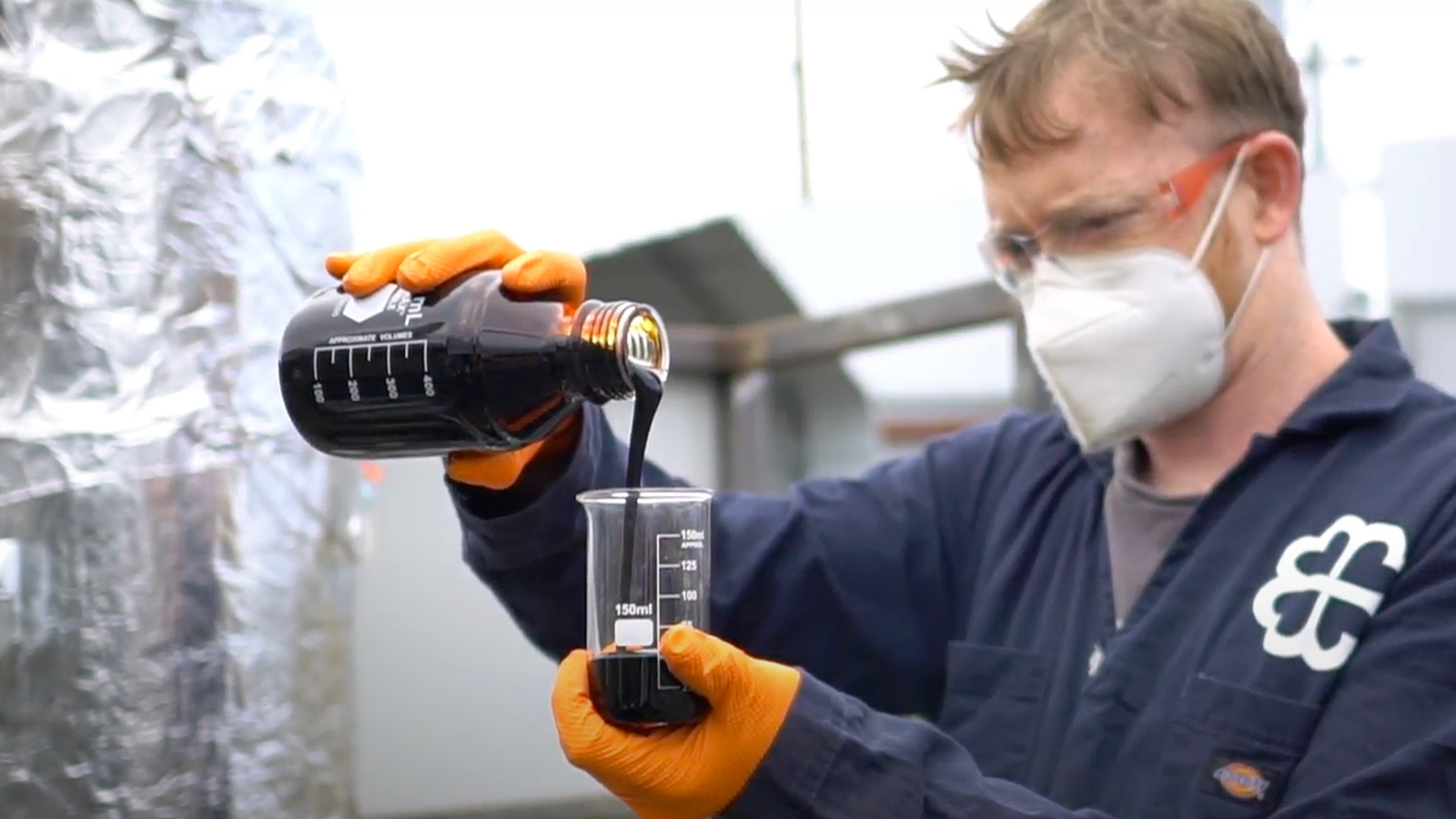 a man in a N-96 mask pours a brown bottle of oil into a beaker