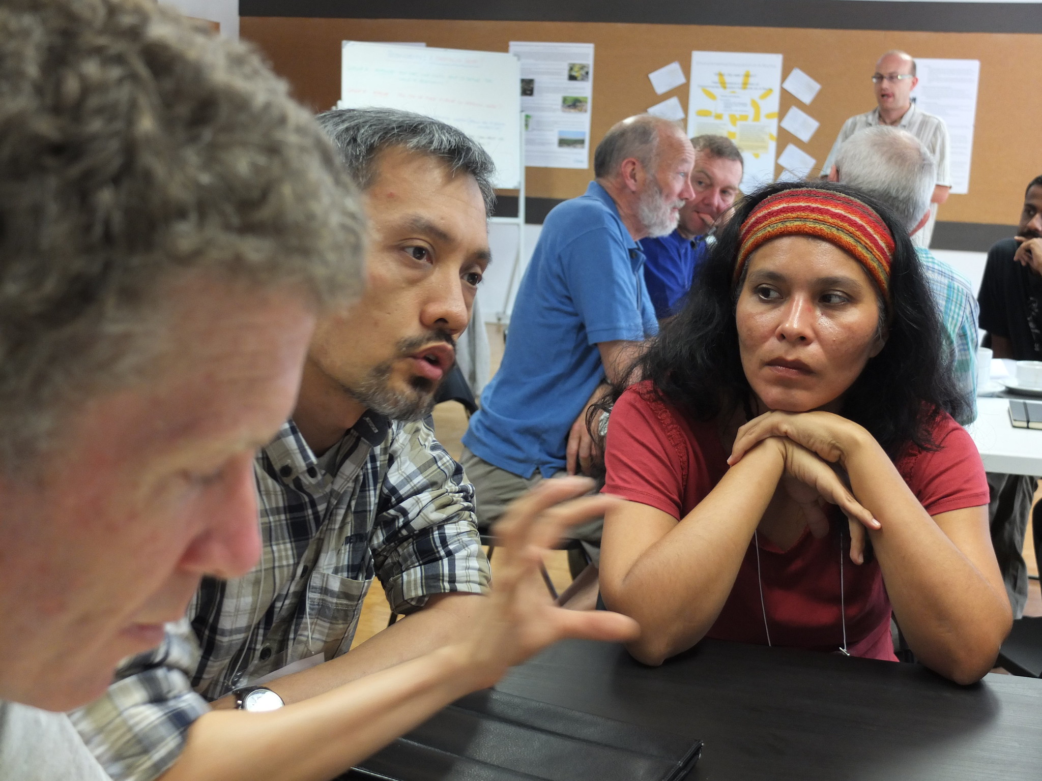 A woman in a red headband and shirt holds her hands under her chin while leaning forward on a table. She is listening to a man with graying hair and a plaid shirt as he gestures with his hand mid-sentence.