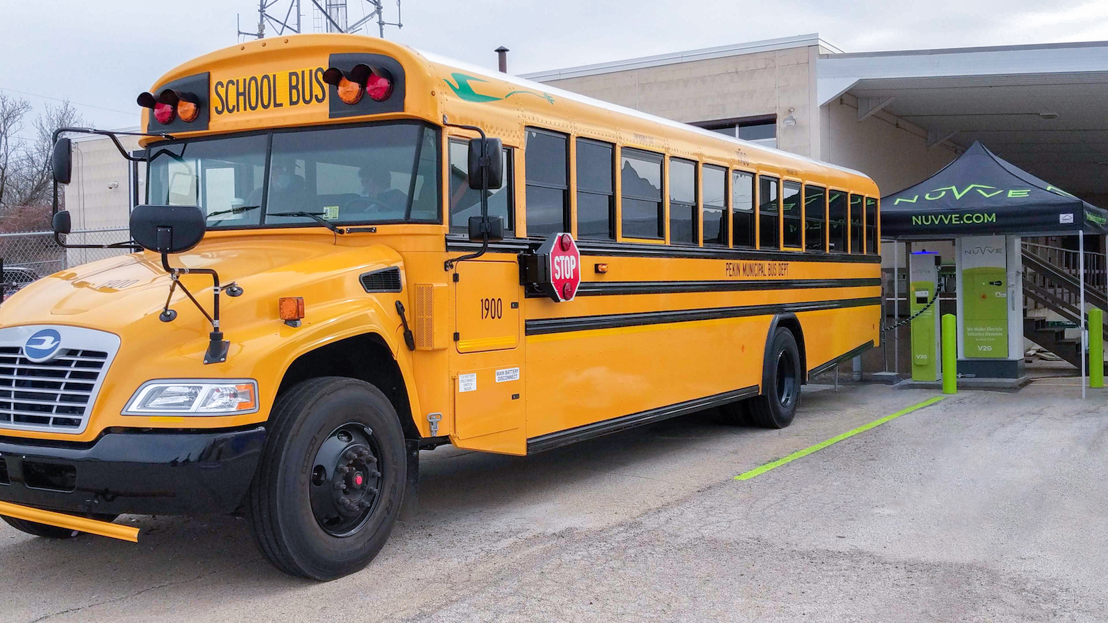 A yellow school bus parked in front of a small outdoor covered charging station labeled "Nuvve"