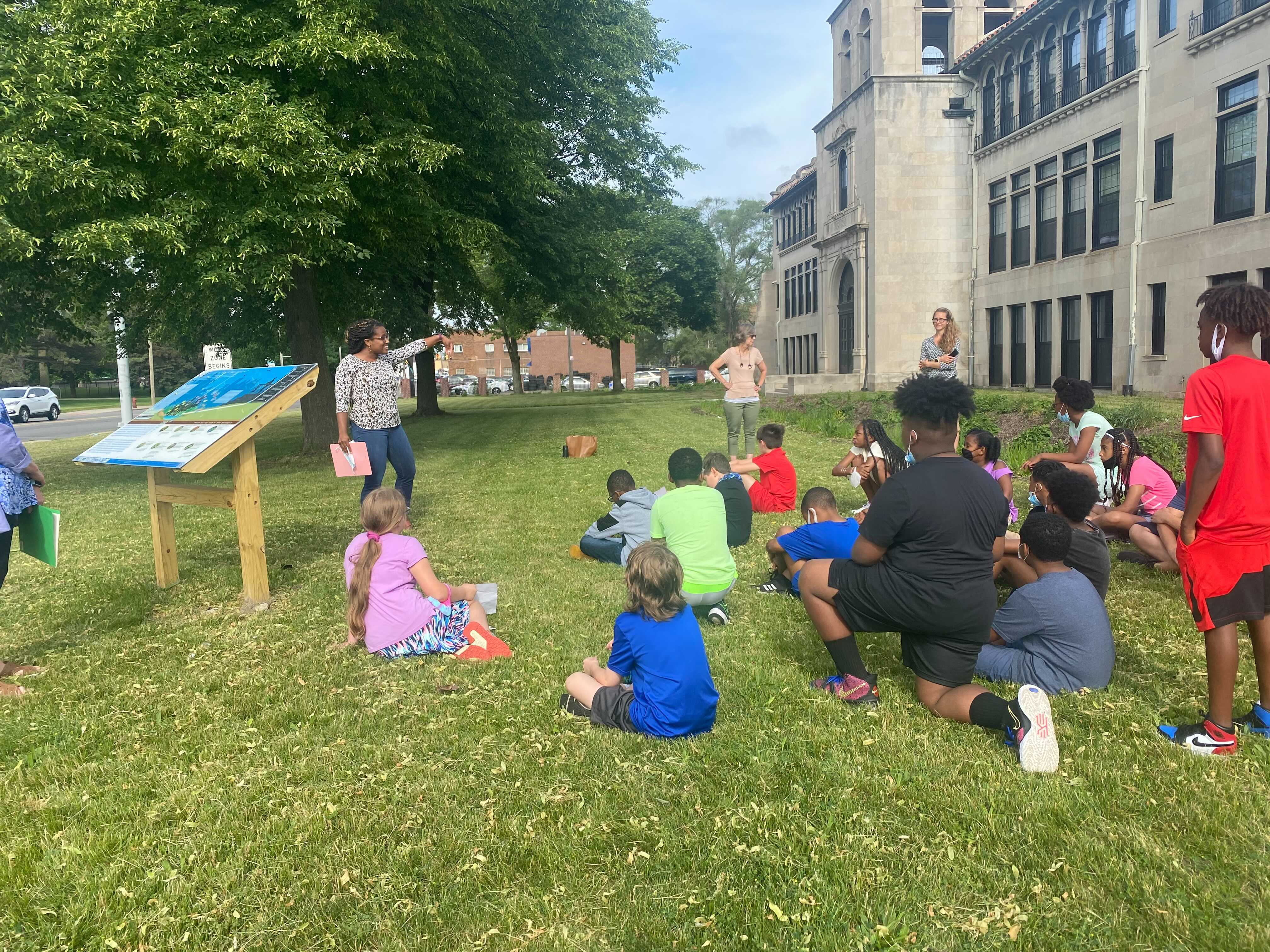 Kids sit on the grass in front of a representative from the National Wildlife Federation as she congratulates them for their work and presents them with a certificate.