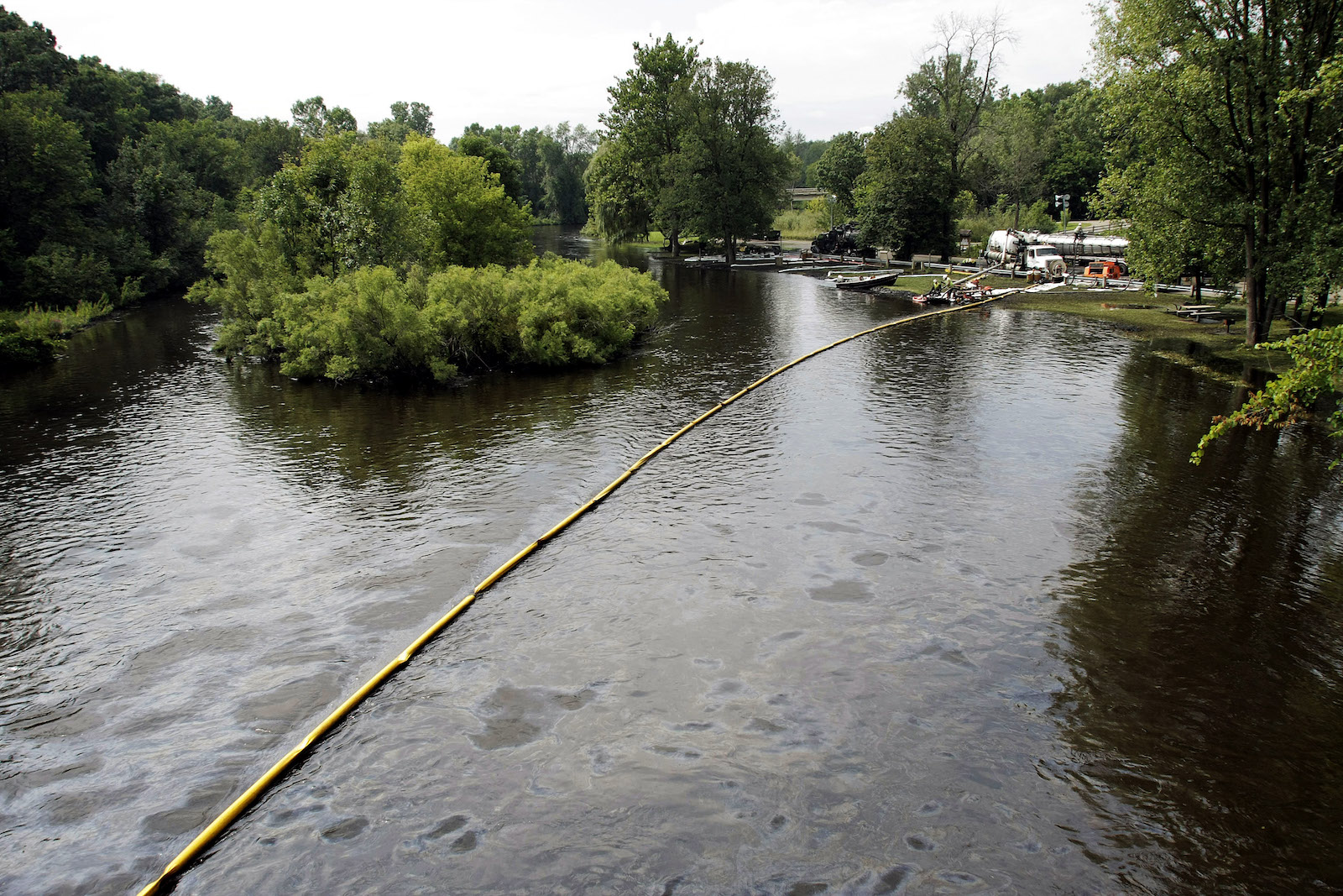 a long pipe cuts across a swath of dark water. In the background, scattered trees and greenery.