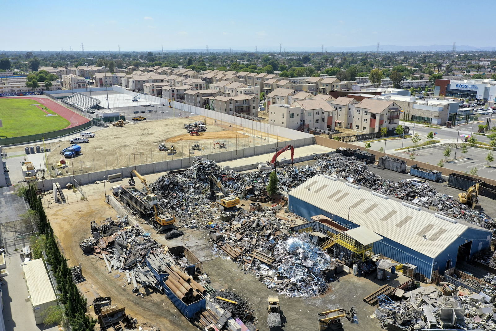 an aerial view of a large scrapyard piled high with metal pieces next to a sunny road and green sports field