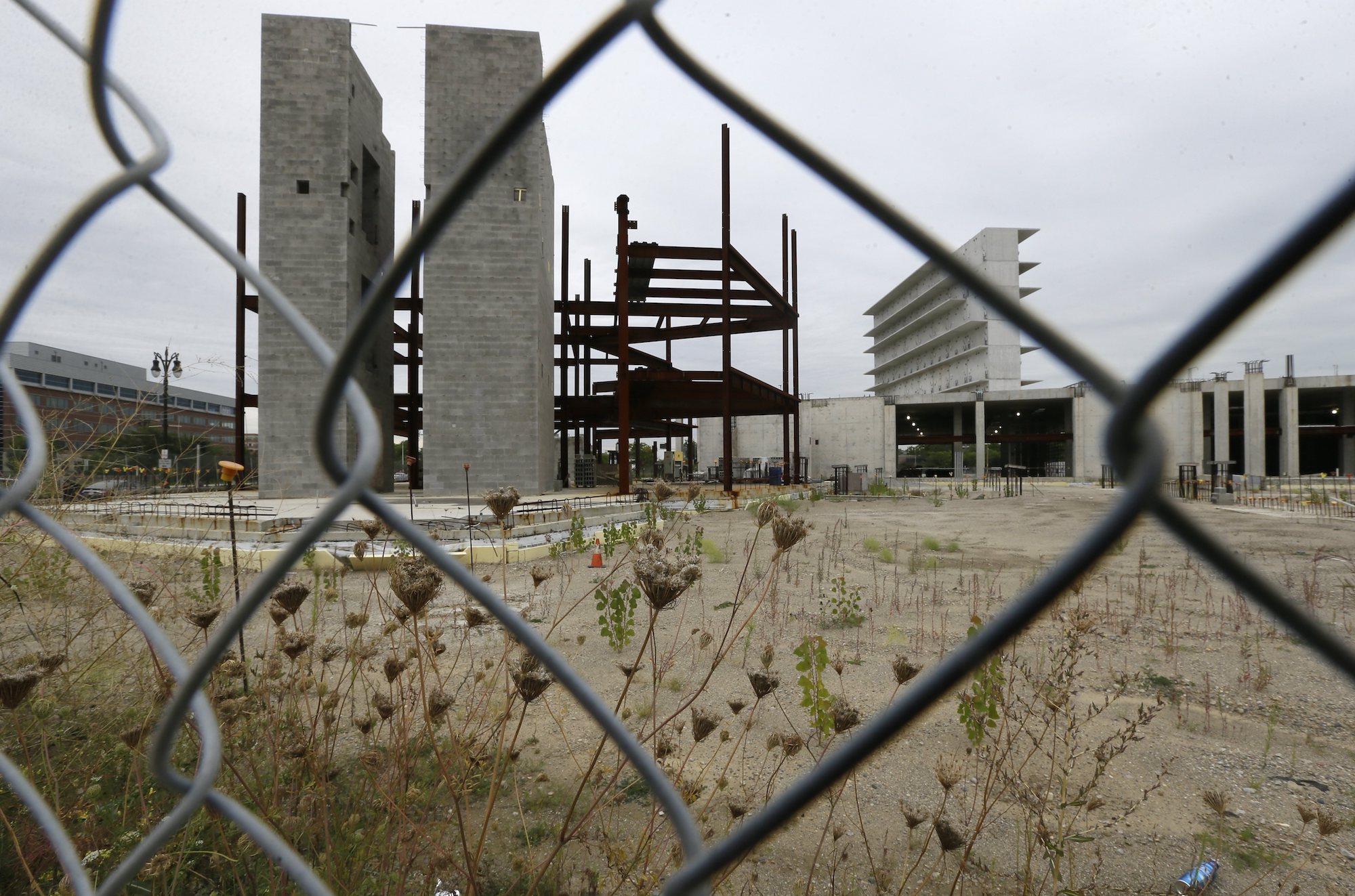 as seen through a fence, a construction site with a pair of tall concrete pillars and a lot overgrown with weeds