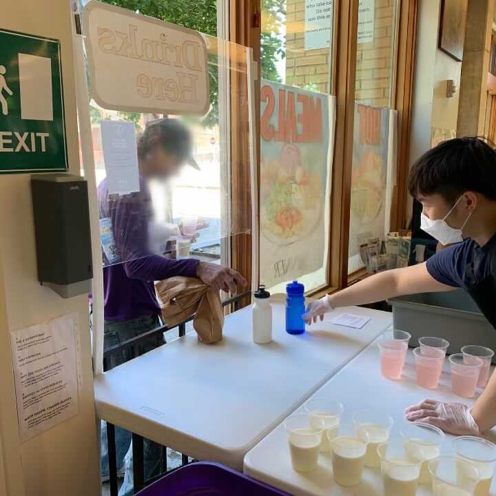 a man in a white mask holds his hand out toward two reusable bottles and a row of drink cups with pink and white liquid. Through a protected walk-up window, another person reaches through to grab a cup