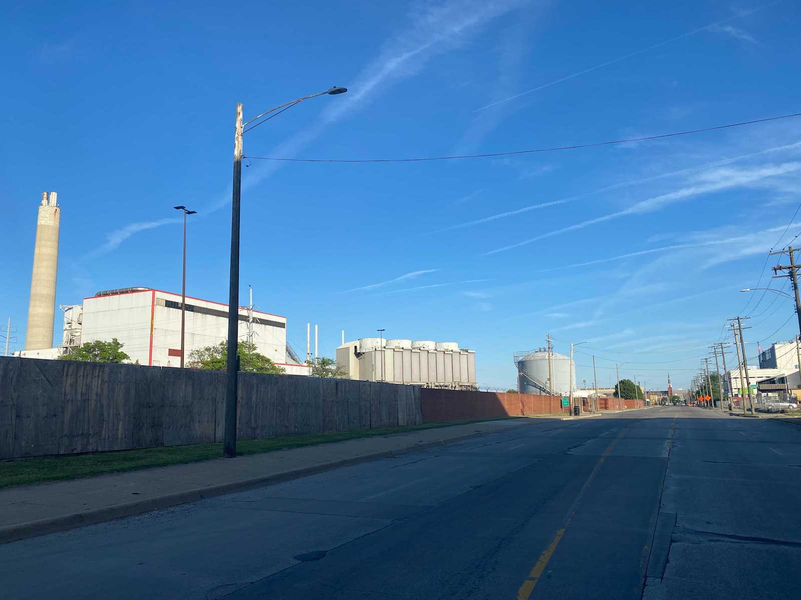 tall concrete structures can be seen behind a green industrial fence alongside a shaded road