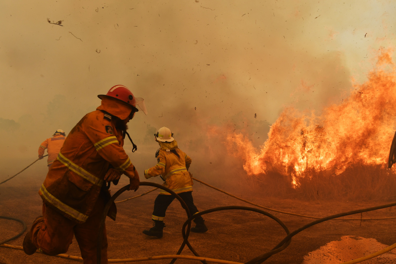 firefighters in full protective gear lift hoses as a fire rages behind them