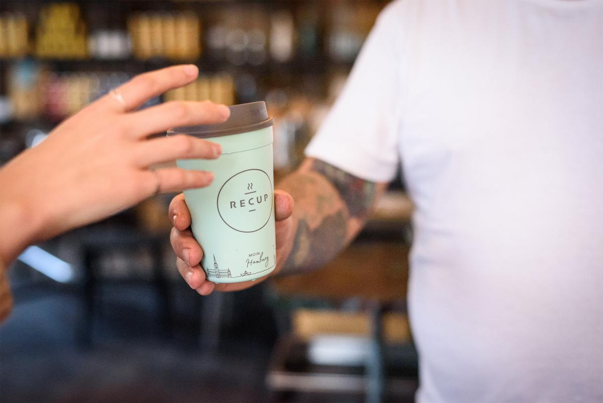 A person reaches for a green coffee cup with brown lid. the cup is labeled ReCUP