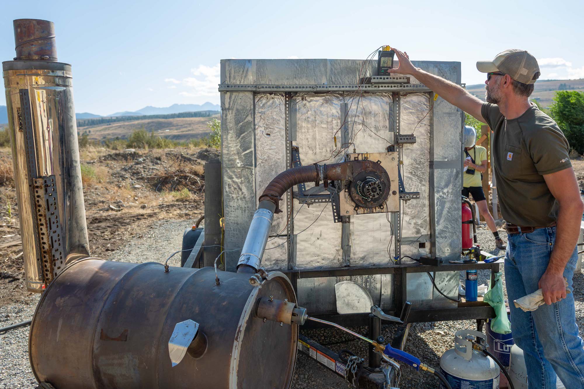 A man wearing a grey shirt and blue jeans holds a measuring instrument on his left hand to check a gauge on a machine called a pyrolyzer, which looks like big, metal garbage container connected to a rusty metal barrel. He's outside, and the sky is blue.