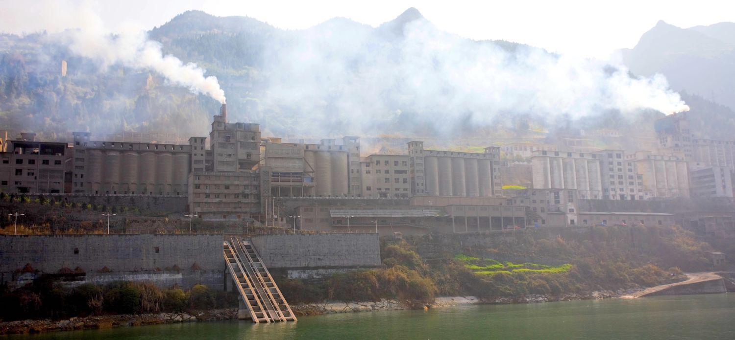 A grey, almost castle-like -looking structure emits smog. Bhind,a mountain range emerges like a wall. In front of it, near to the lens, a green, calm river passes by. A few small trees, with red and green foliage, indicate that the photo was taken during the fall months.