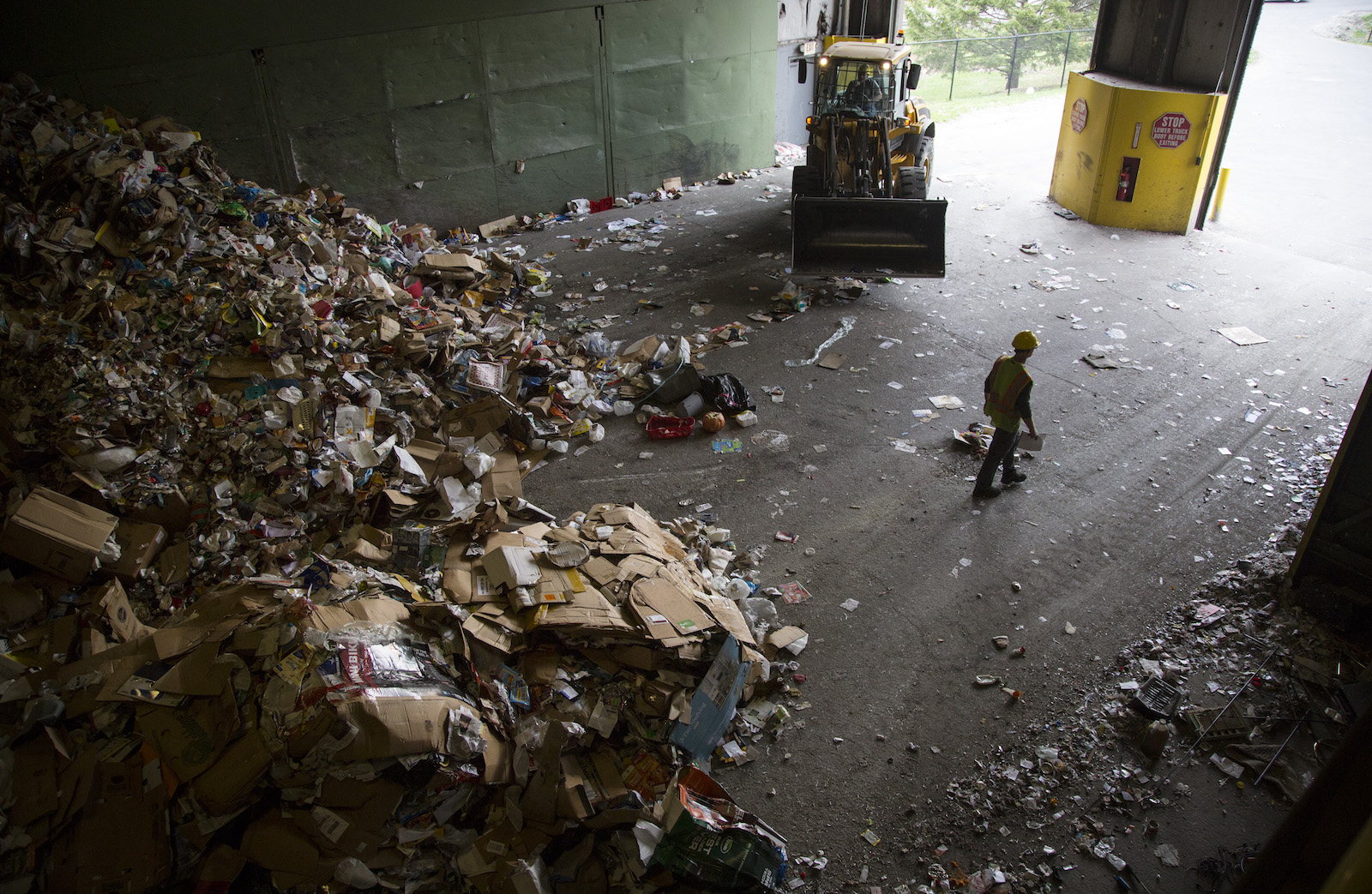 a large pile of boxes and bottles sits inside a warehouse, dwarfing a worker who is walking by