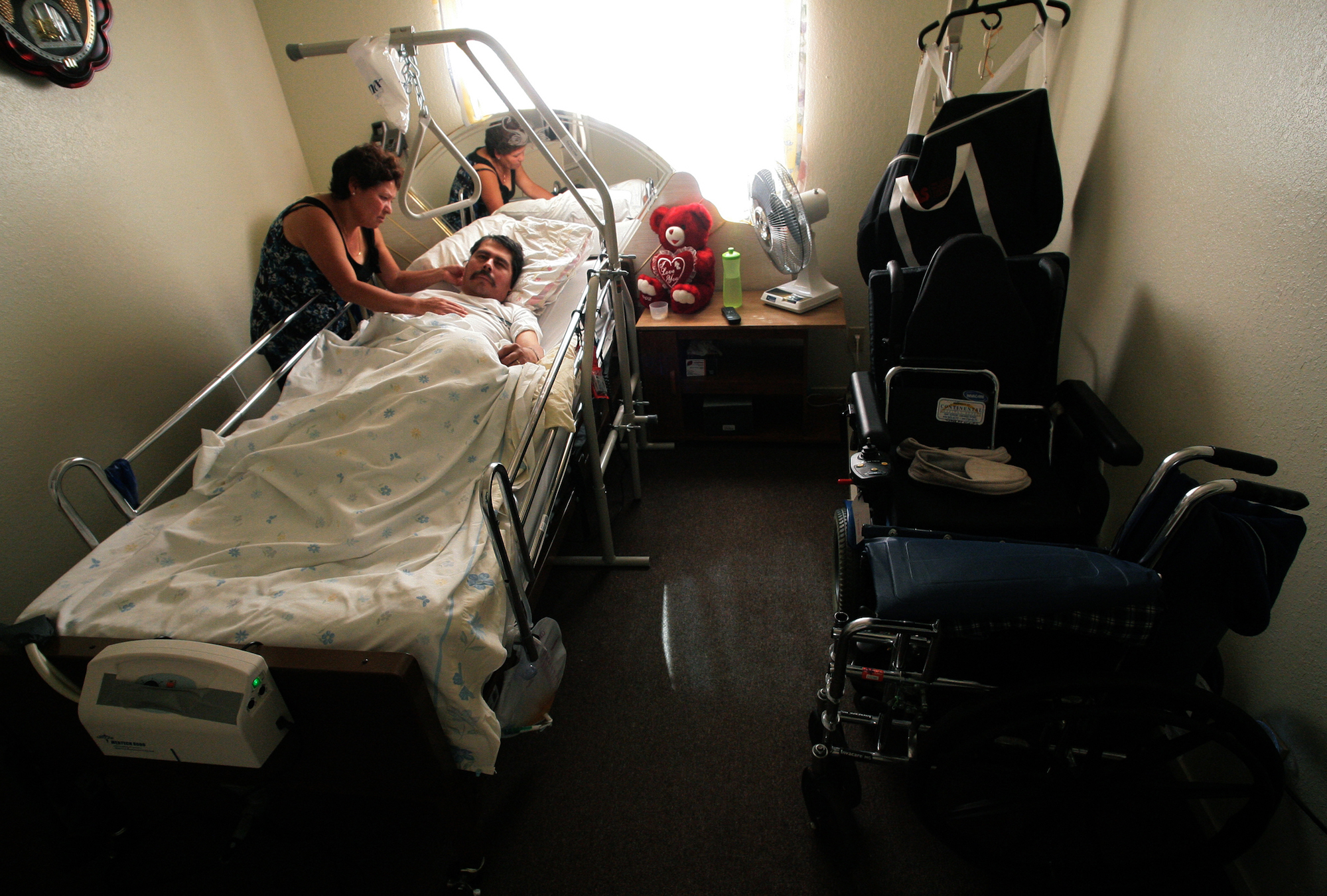 a woman leans over a hospital bed with a man lying in it. The bed is in a small room situated near a window