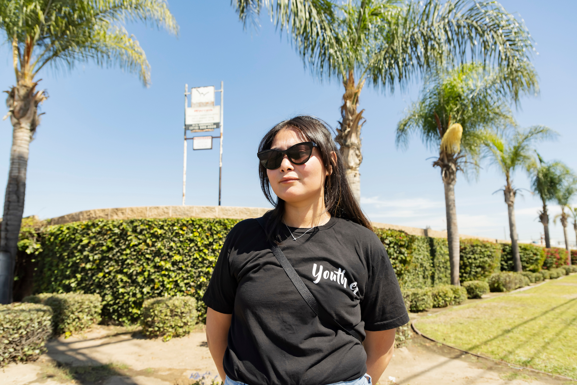 a woman with long black hair and sunglasses stands in front of a sunny street with oil infrastructure in the background