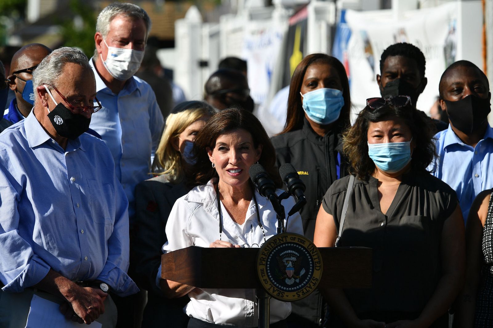 New York Governor Kathy Hochul is standing in he middle of a street in Queens, talking through. microphone and accompanied by New York City's mayor Bill de Blasio and her team