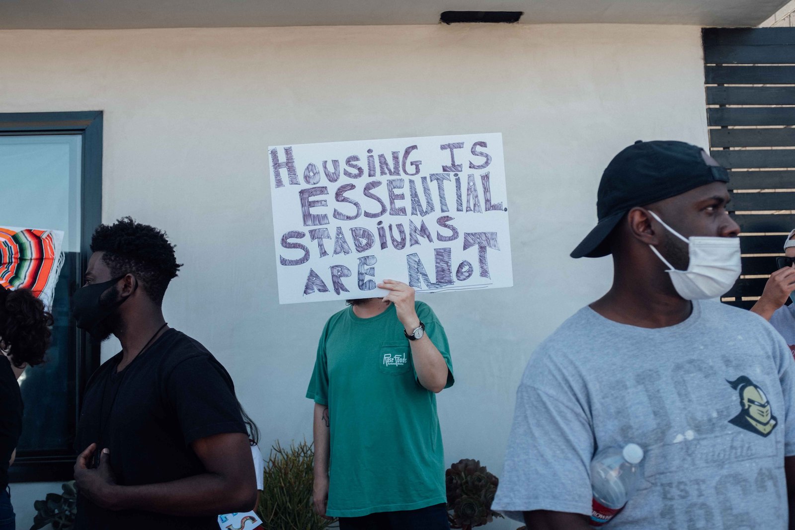 a person in a green t-shirt holds a sign that says housing is essntial stadiums are not