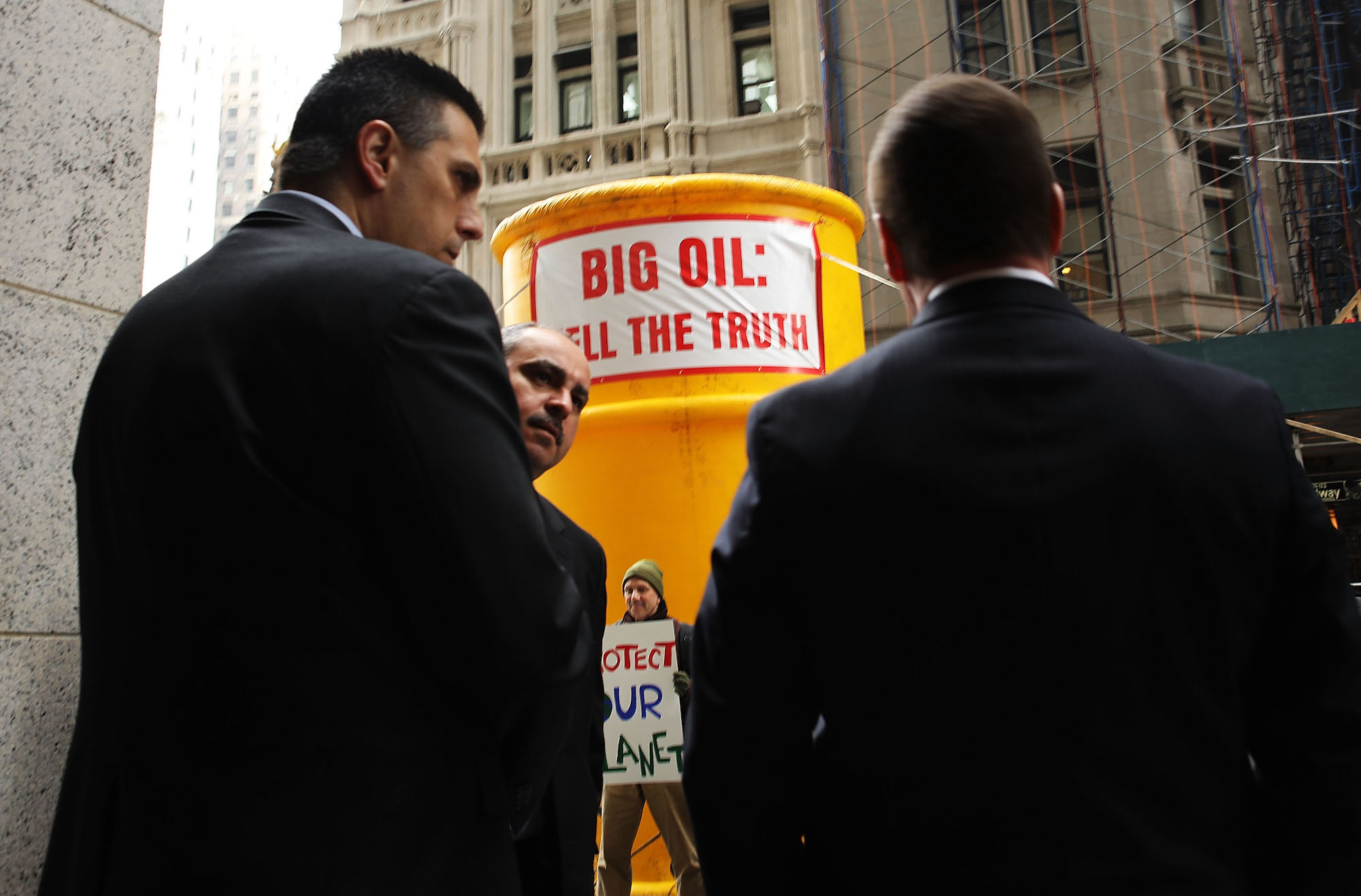 A canary-yellow blow-up stands in front of New York State Attorney General's office. Protestors rally around it, while two security guards watch the scene.