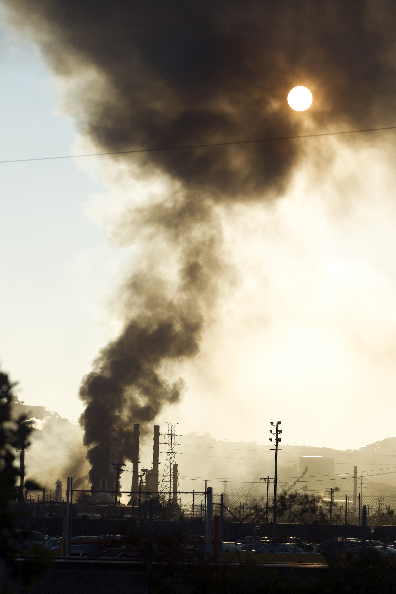 a large cloud of smoke from a silhouetted refinery reaches high into the sky partially obscuring the sun