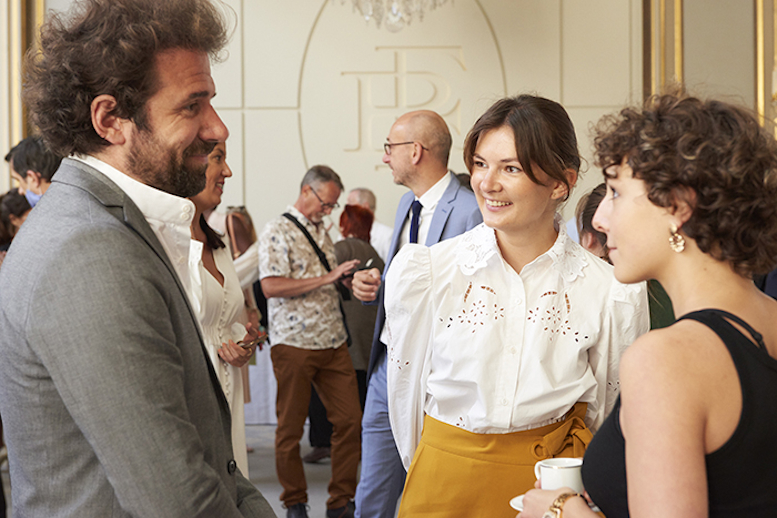 a woman in a white lace shirt and yellow skirt smiles in a group conversation with a man in a gray jacket and white shirt and a woman in a black short-sleeved shirt and gold earrings