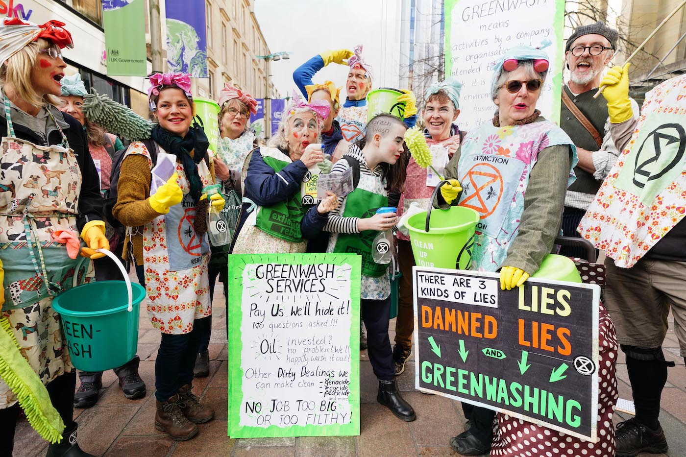 Protesters dressed in aprons and cleaning gloves holding buckets with signs that say greenwash services