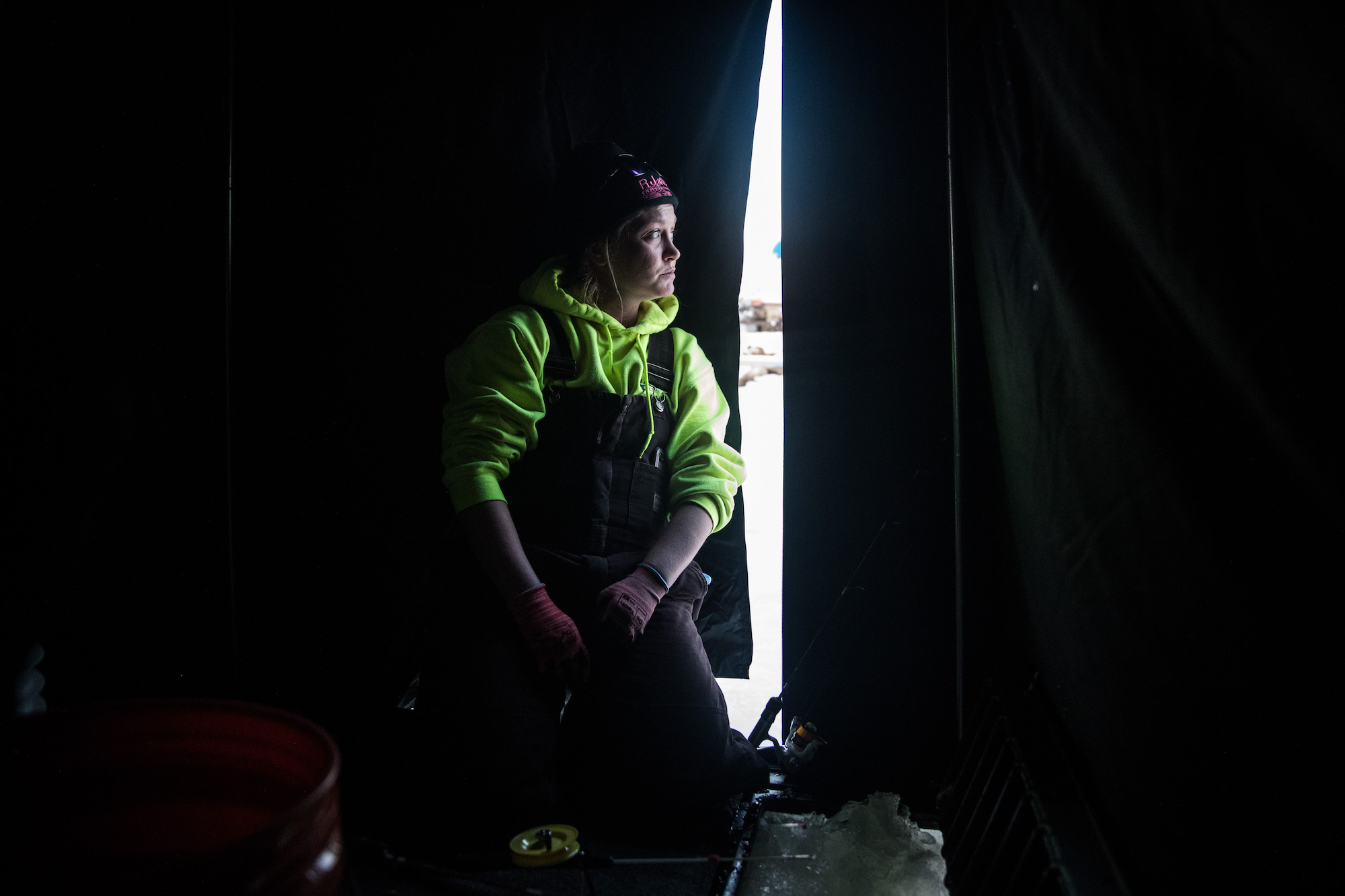 A blonde woman in a black hat, green sweatshirt, and black snow overalls peeks out of a glowing, white slit in an a dark tent. She is seated and there are some fishing implements visible near her legs.