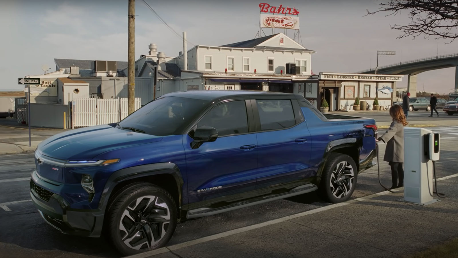 Woman charging a royal blue electric Chevy Silverado with seafood restaurant in background