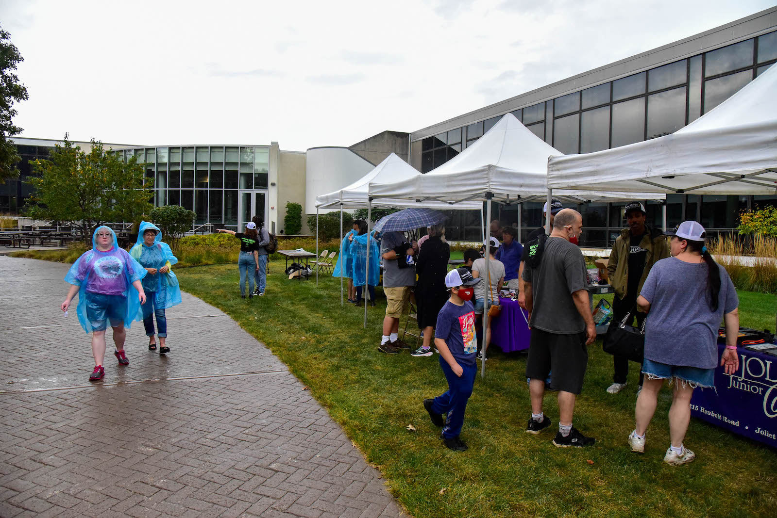 two people wearing translucent blue rain ponchos walk along a road next to a lawn where tents are set up with tables. People stand by the tables with purple tablecloths