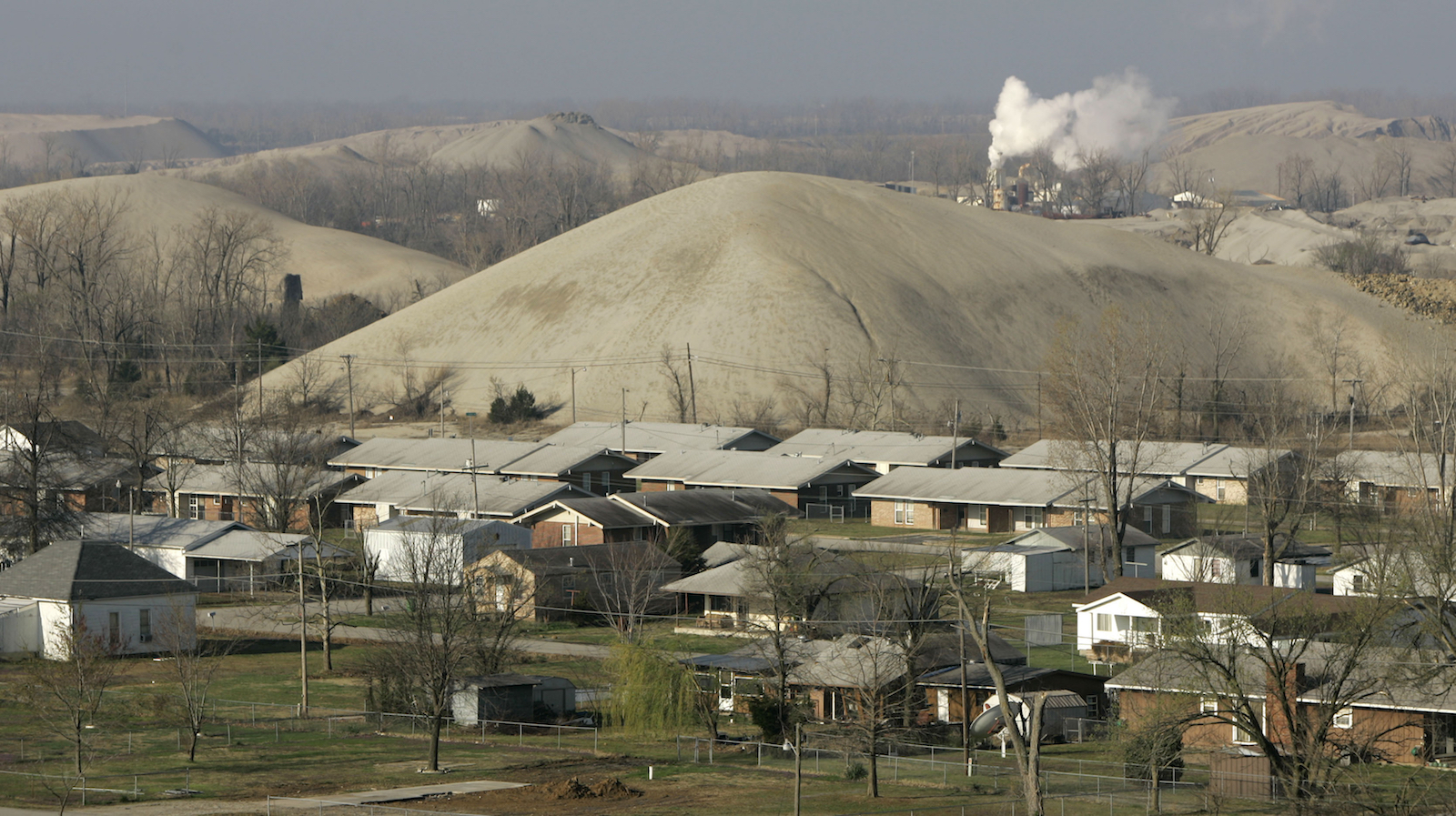 a cluster of a dozen single-story houses with dead-looking trees and large gray hills surrounding the area