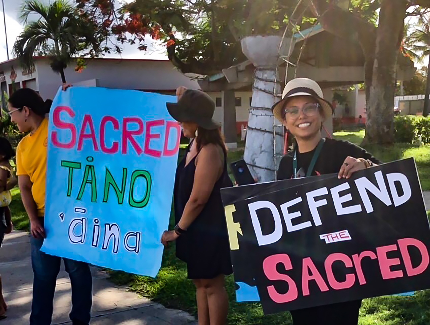 Three people with protest signs reading "Sacred tåno 'āina" and "Defend the Sacred"