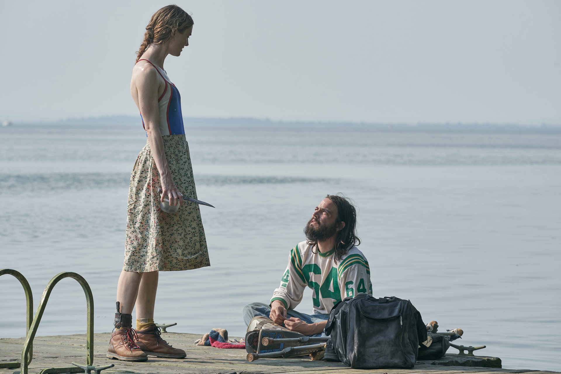 A woman standing, facing a man sitting, on a dock on a lake