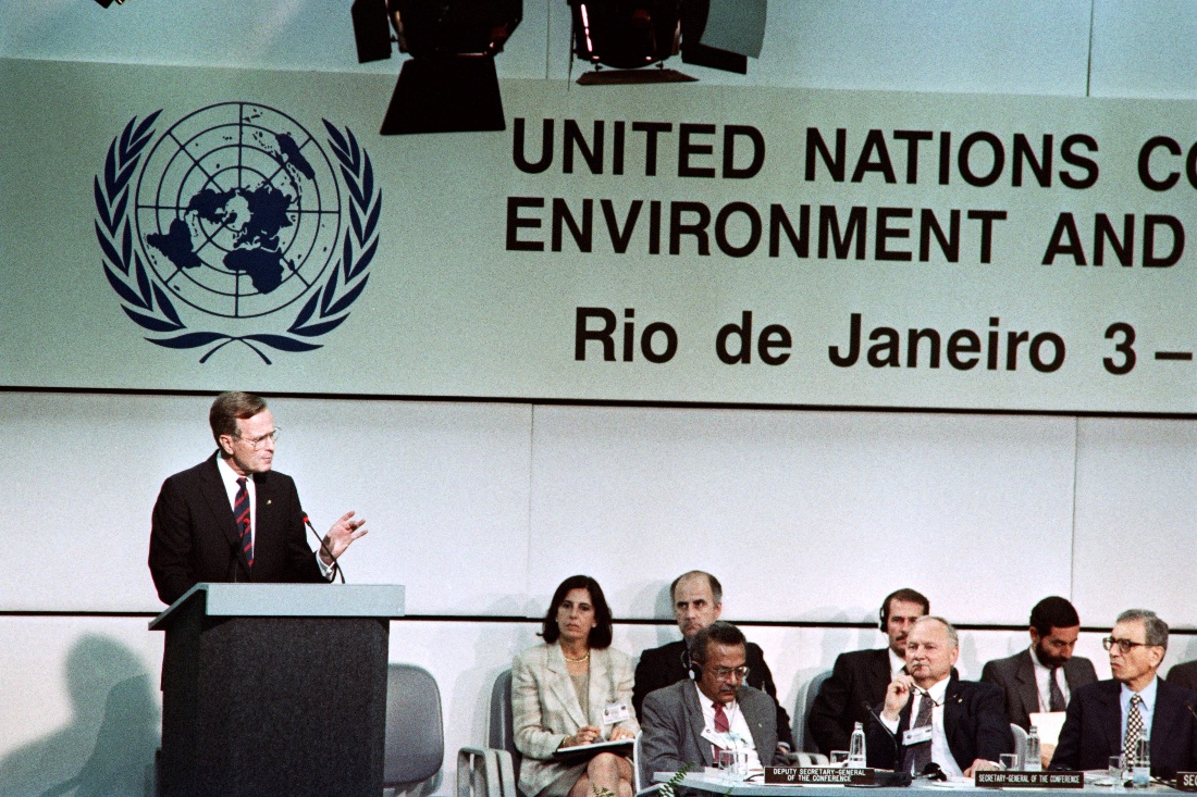 Former President George H W Bush speaks at a podium next to seated officials.