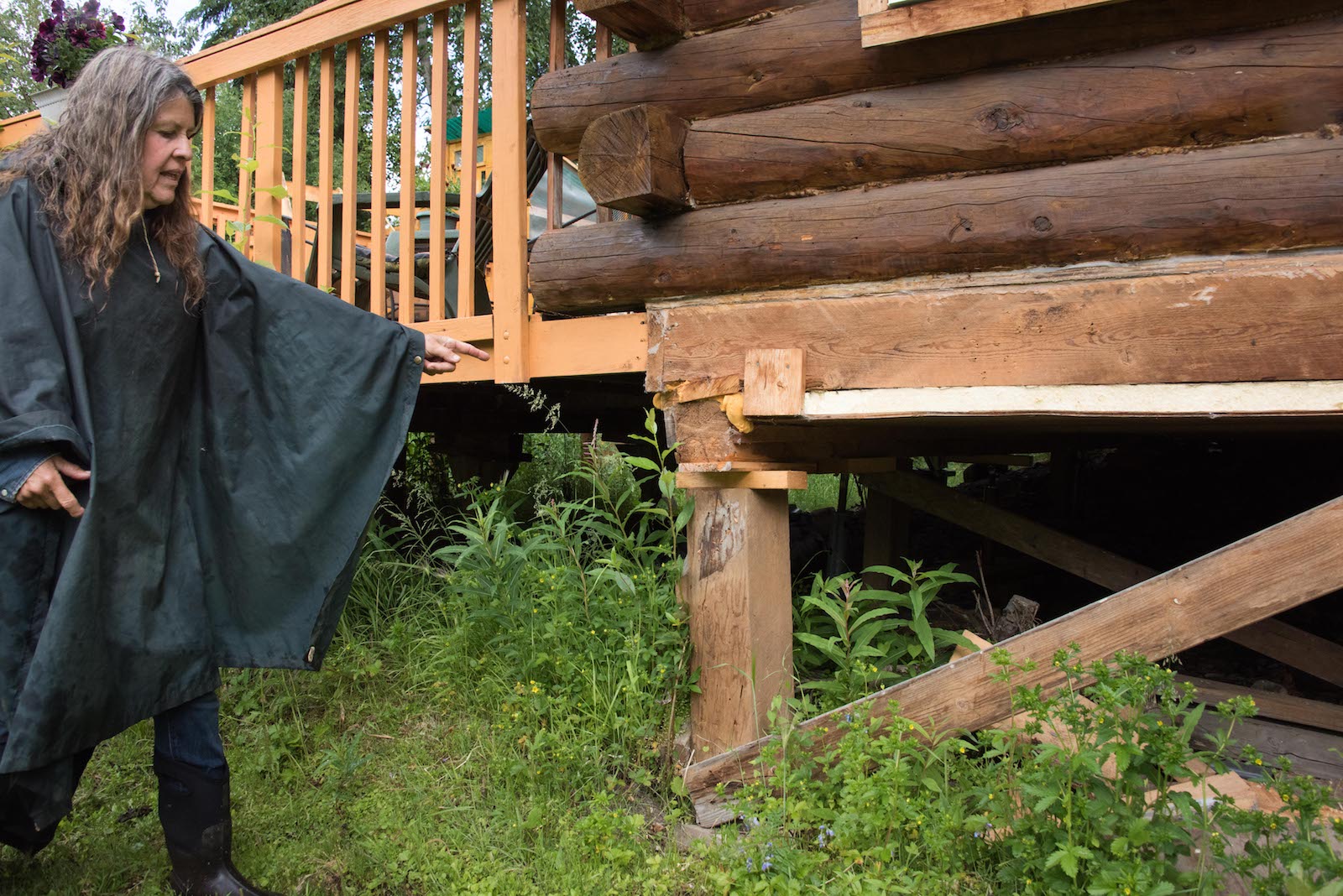 a woman in a green poncho points to a wooden support on the corner of a wooden house