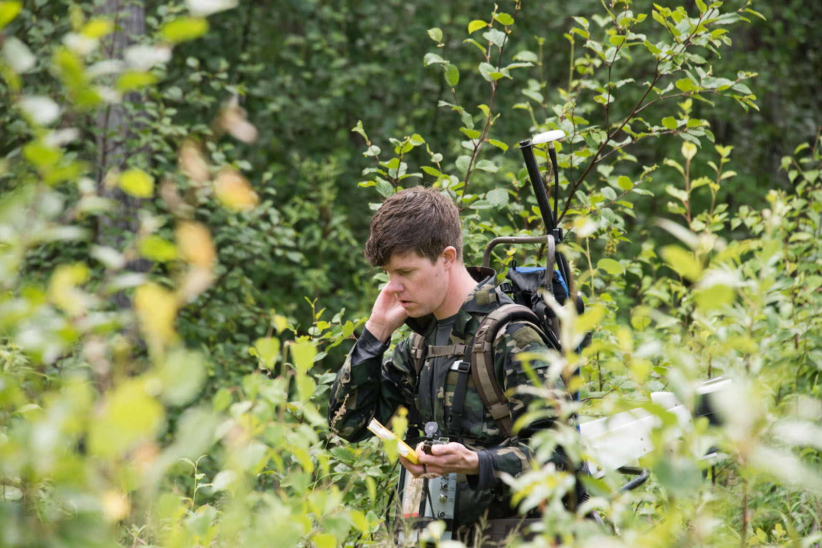 a man in green camoflague fatiges holds his hand to his ear while standing in a green lush area