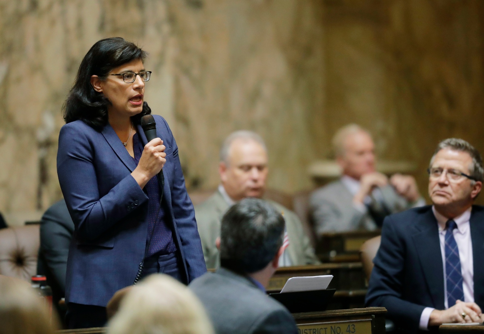 A woman in a blue blazer speaks into a microphone as others watch.