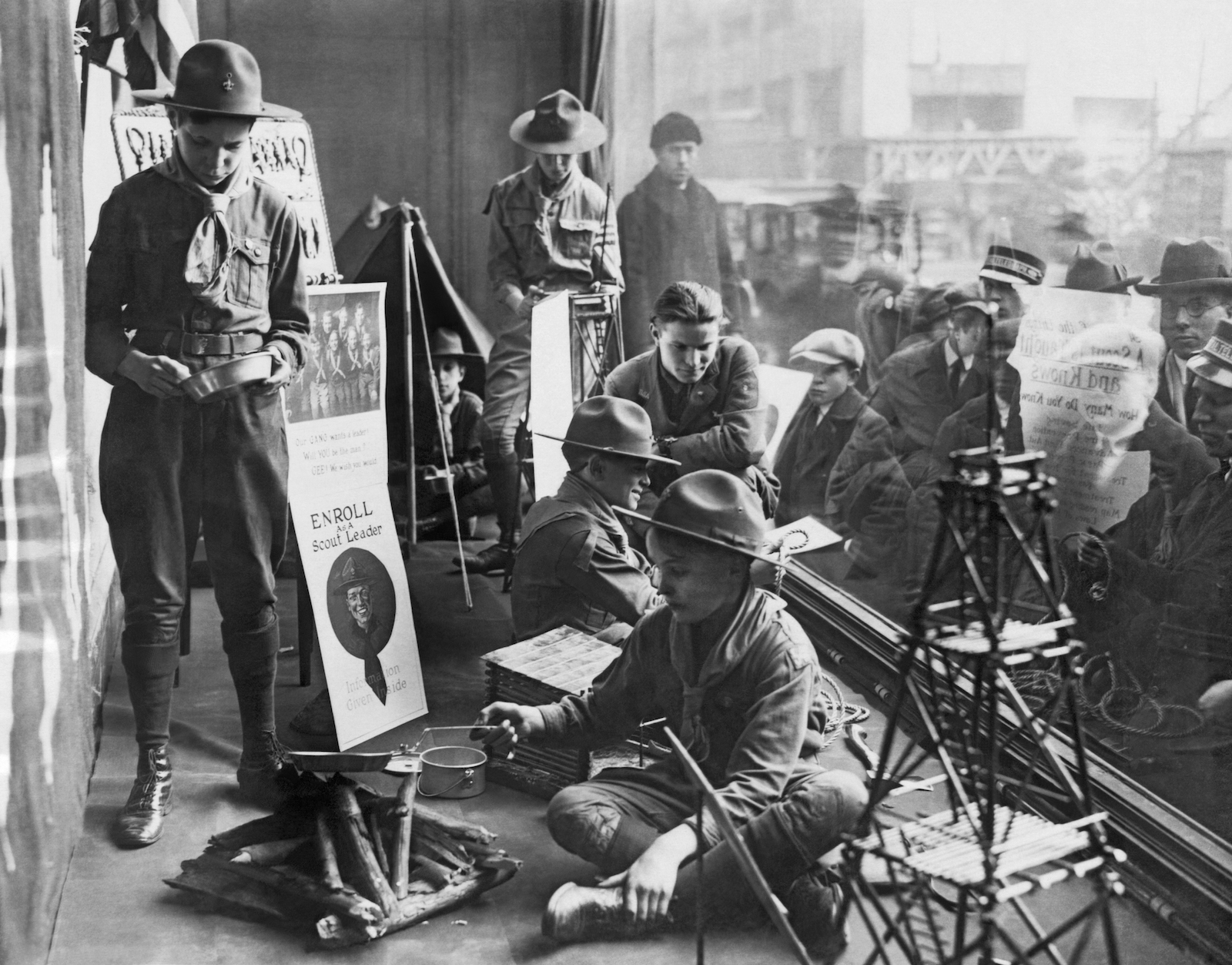 a black and white photo of a group of boys in scout uniforms sitting and standing inside a store near a window