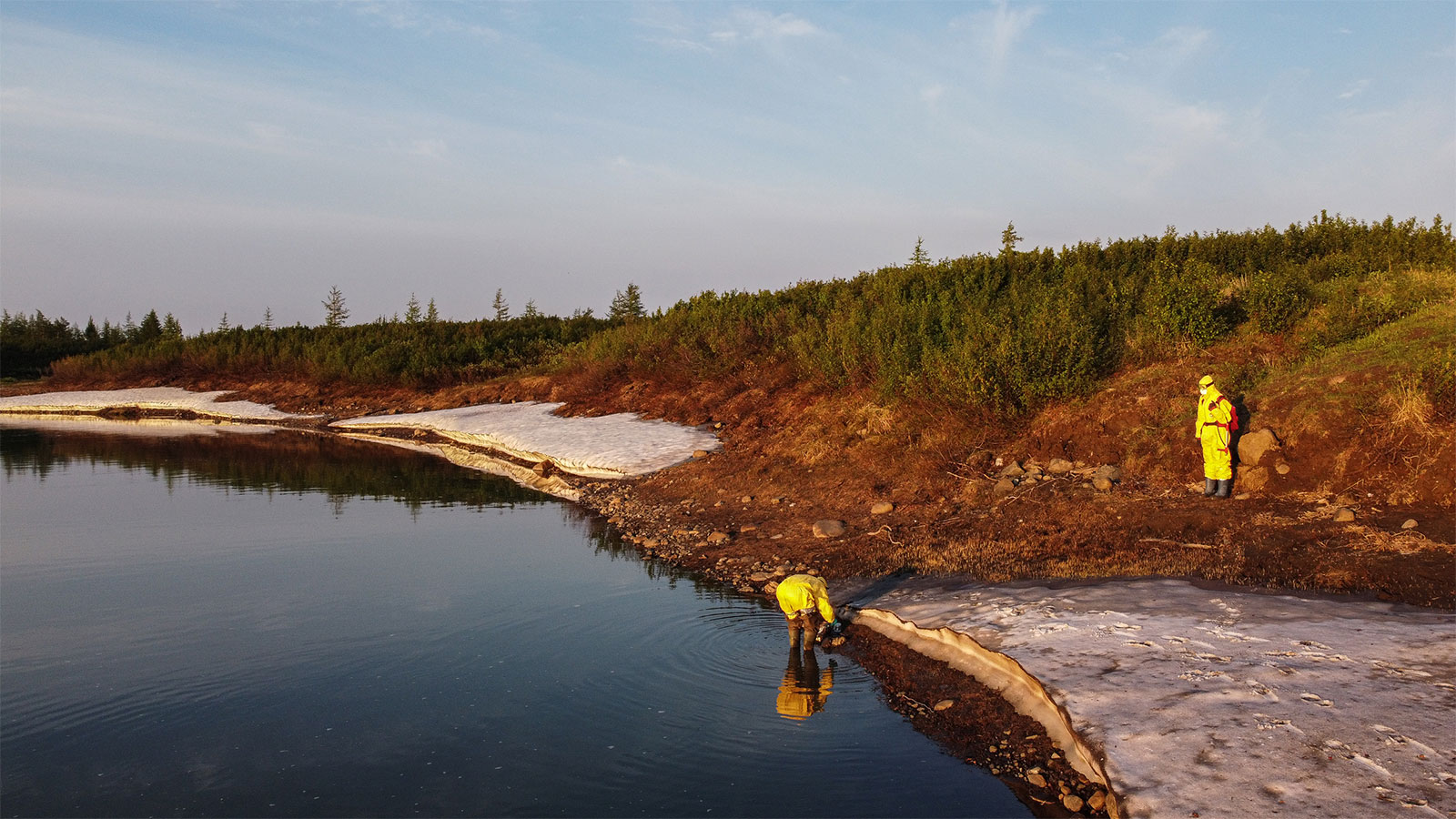 Two people in yellow hazmat suits, one standing on the bank of a river and the other taking a sample of river water