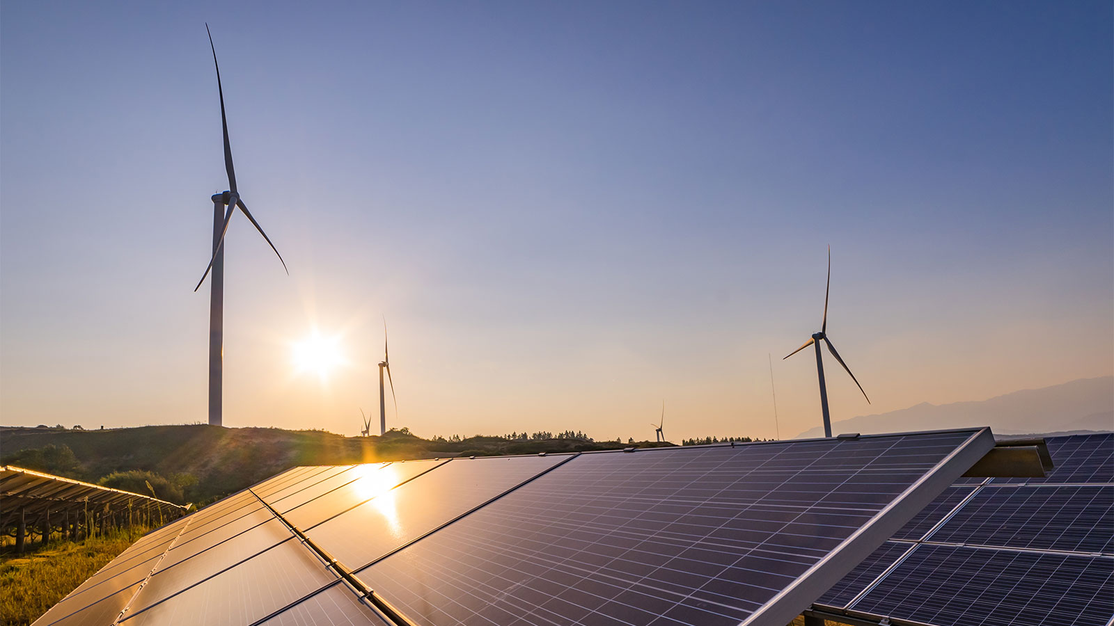 Wind turbines against a blue sky with sun shining low on horizon; solar panels in foreground reflecting the sunlight