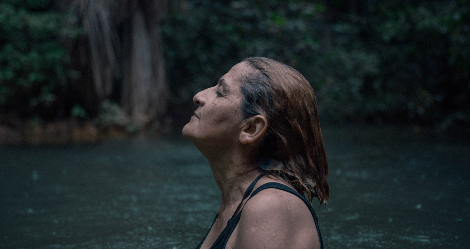 A woman with closed eyes stands in front of a waterfall.