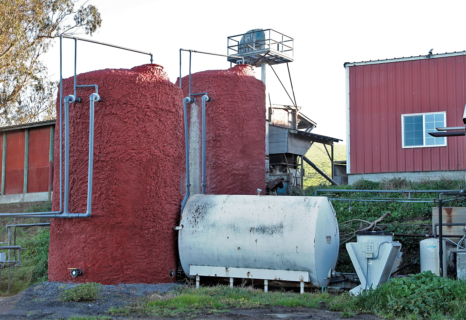 A methane digester on a farm, consisting of two tall red tanks and one shorter gray tank with red buildings in the background