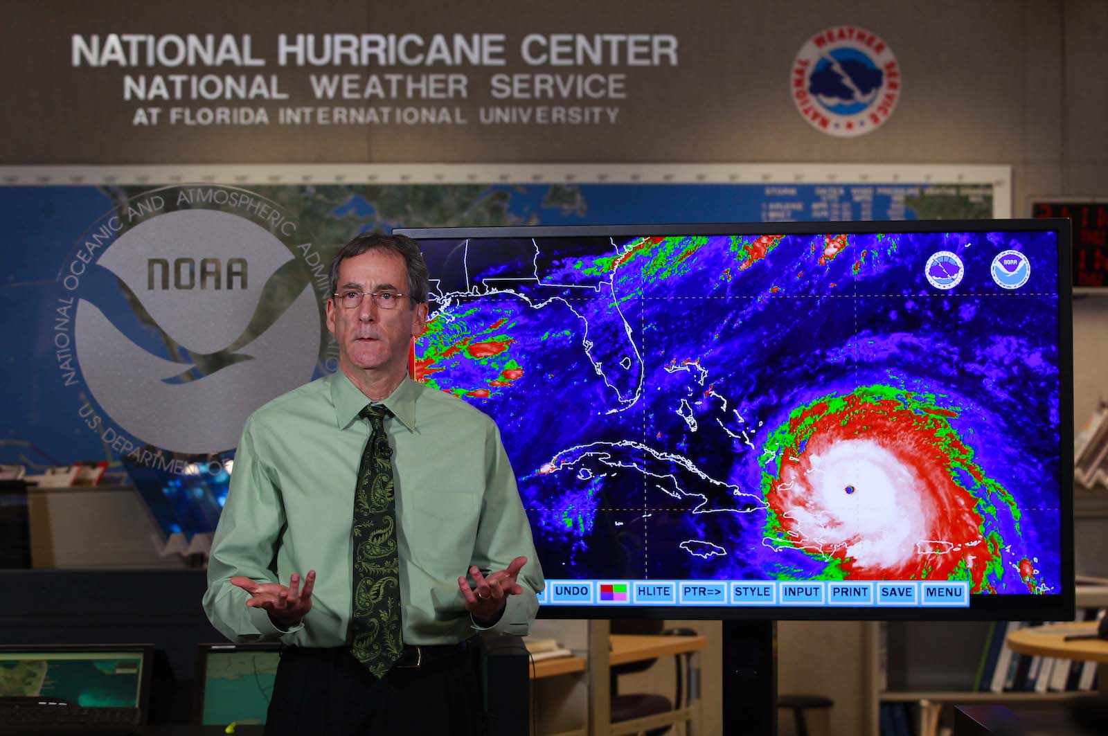 a man in a tie stnads in front of a weather screen for the national weather servie