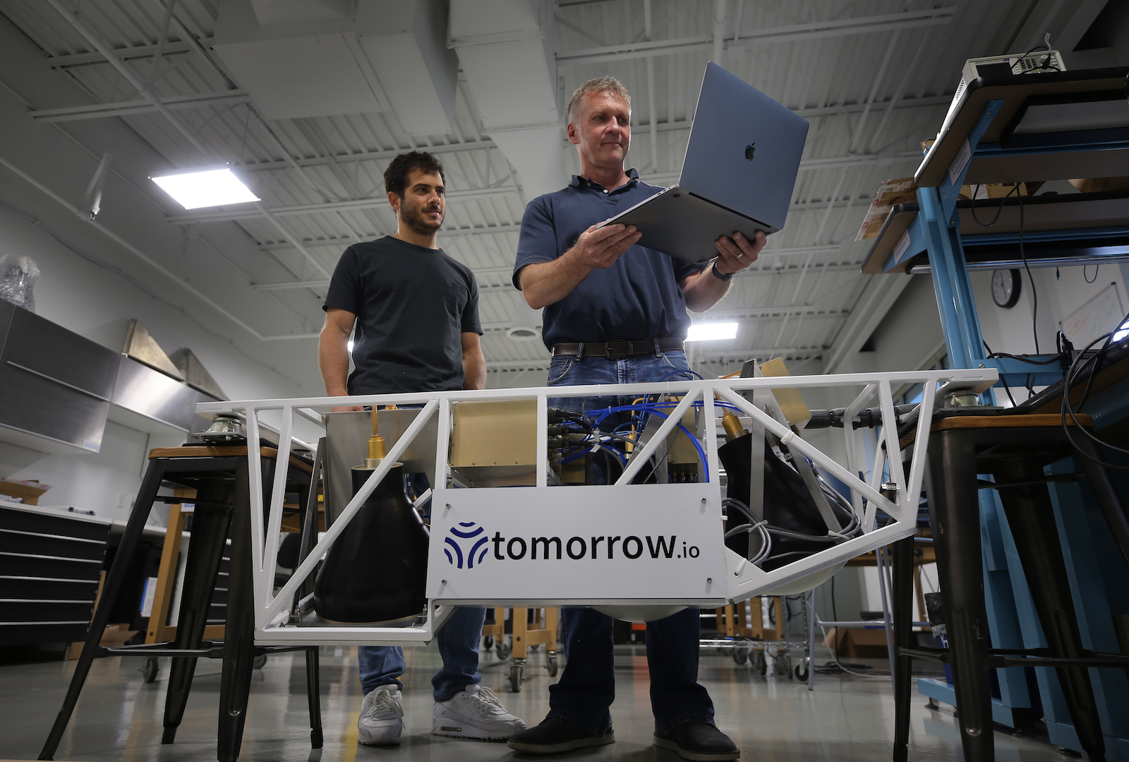 two men stand in front of a gate labeled tomorro.io and look at a laptop