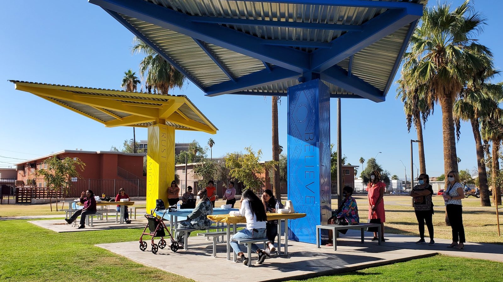 people sit at picnic tables under a shaded pavilion in a park