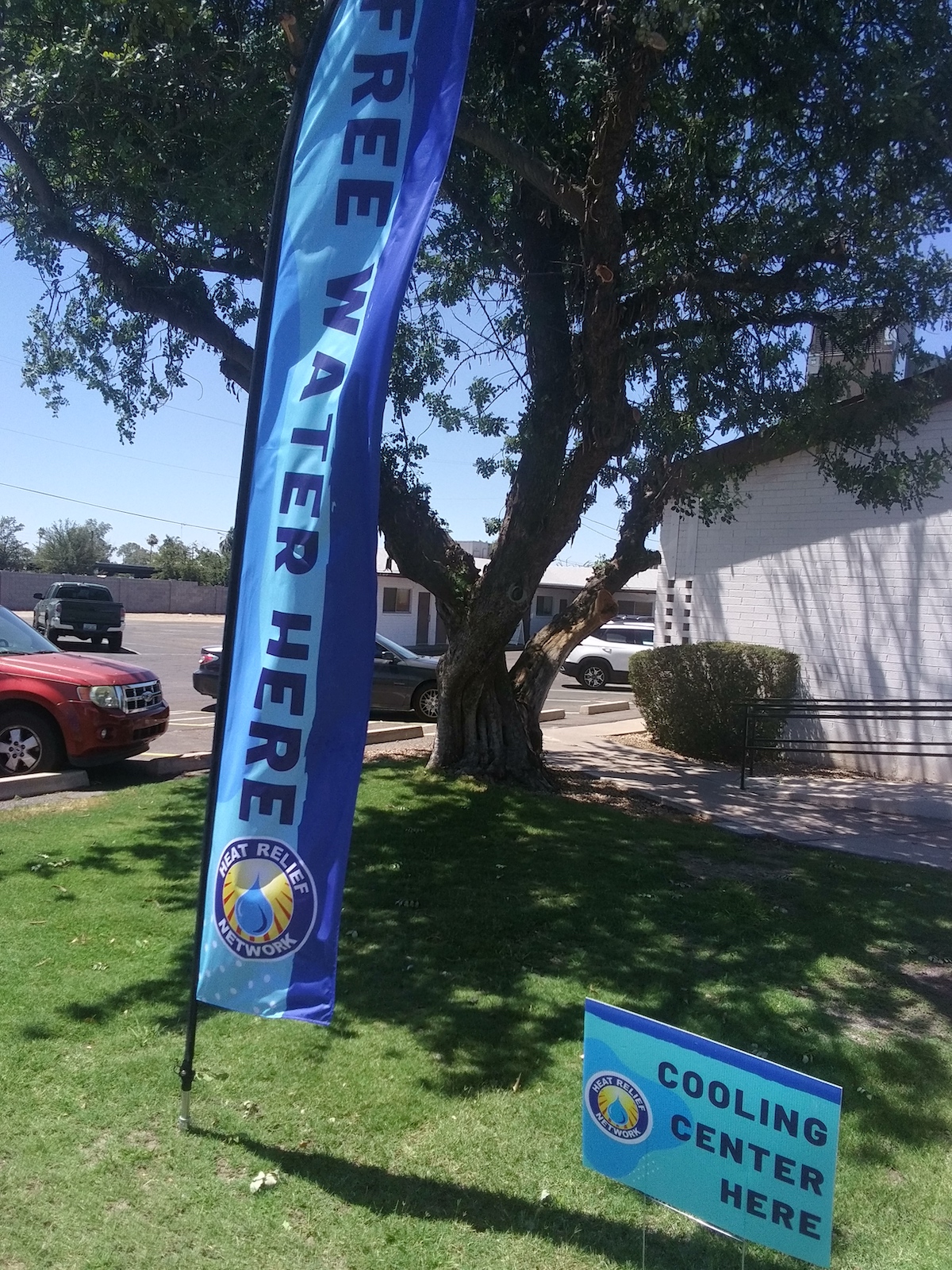 signs reading "free water here" and "cooling center here" in front of a tree and white building