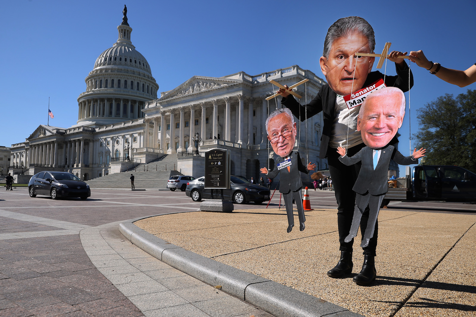 a protester wearing a joe manchin cutout mask holds puppets dressed like Chuck Schumer and Joe Biden