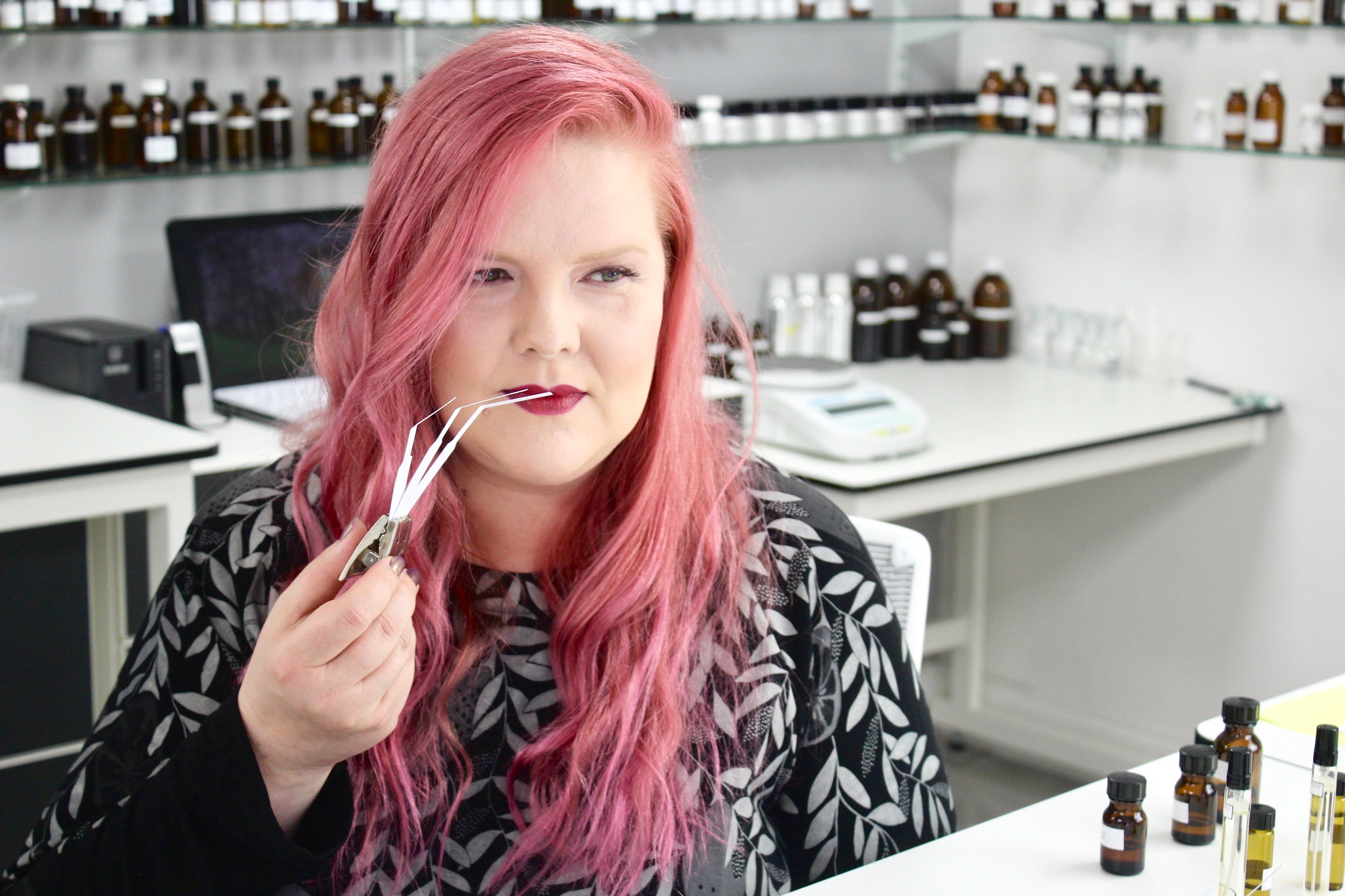 a woman with long red hair holds an eyedropper up to her nose