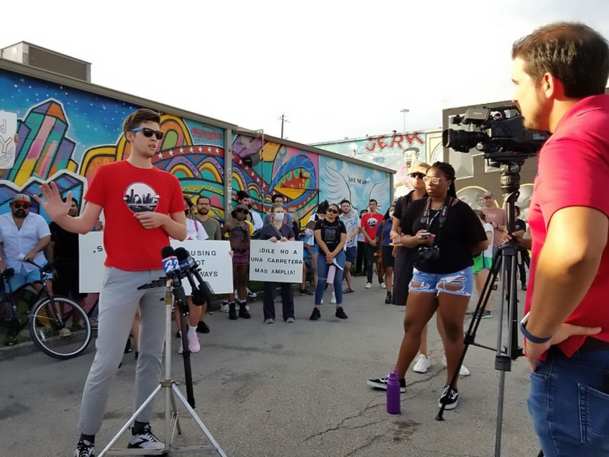 a man in a red shirt stands in front of a camera flanked by people holding signs