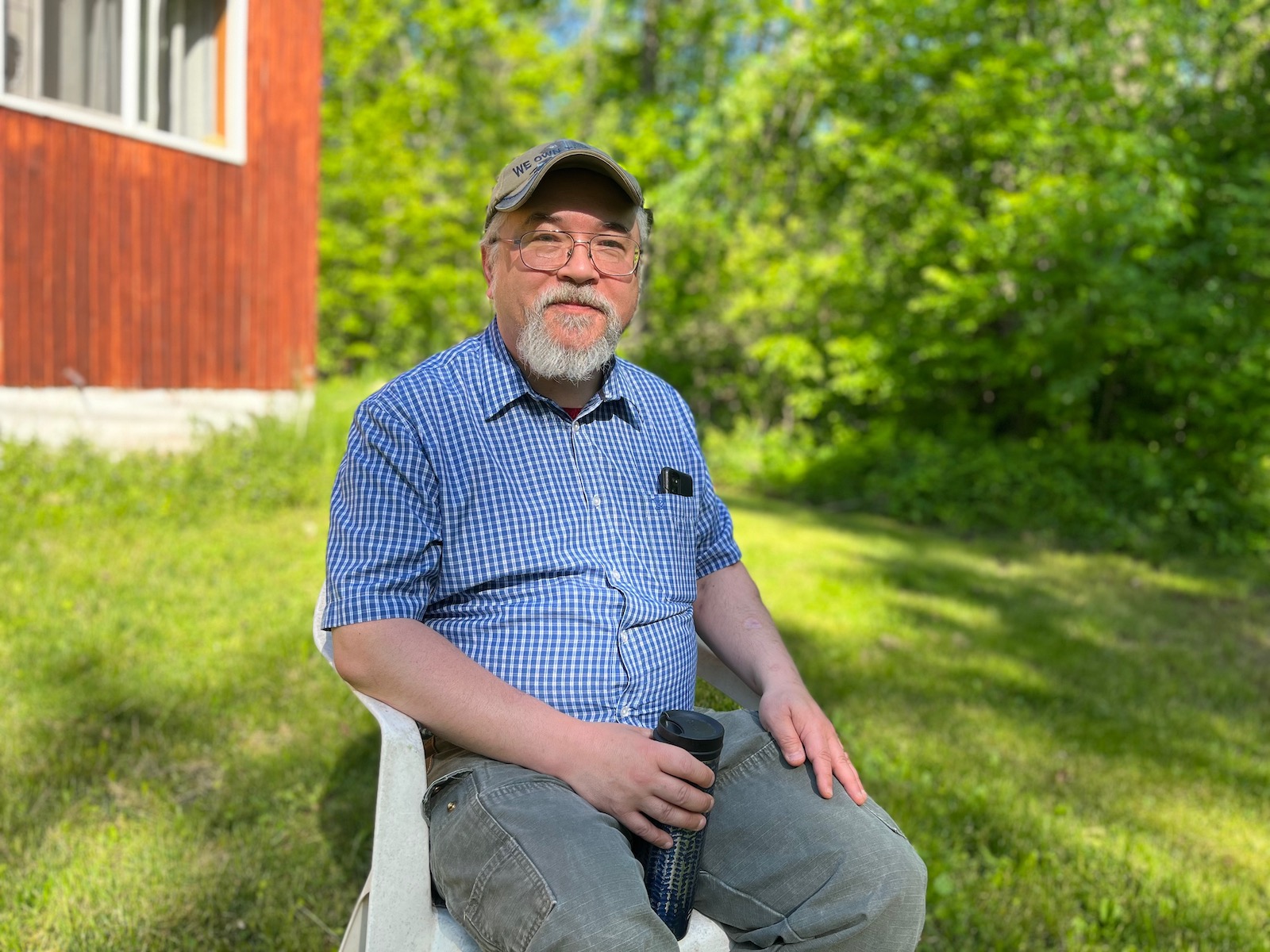 A man in a blue shirt and baseball cap sits in front of a red house