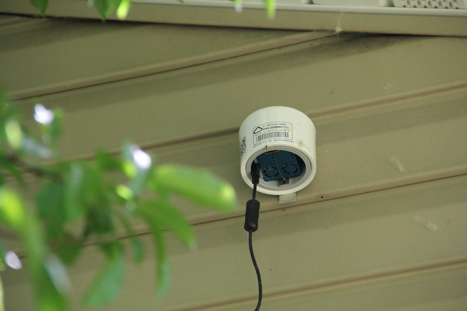 a small white canister with a black cord hangs outside a house with leaves in front of it